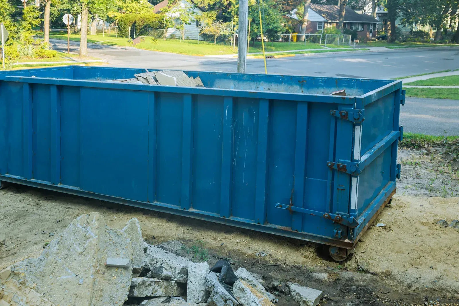 Blue construction dumpster with rubble, parked on a driveway, street and trees in the background.