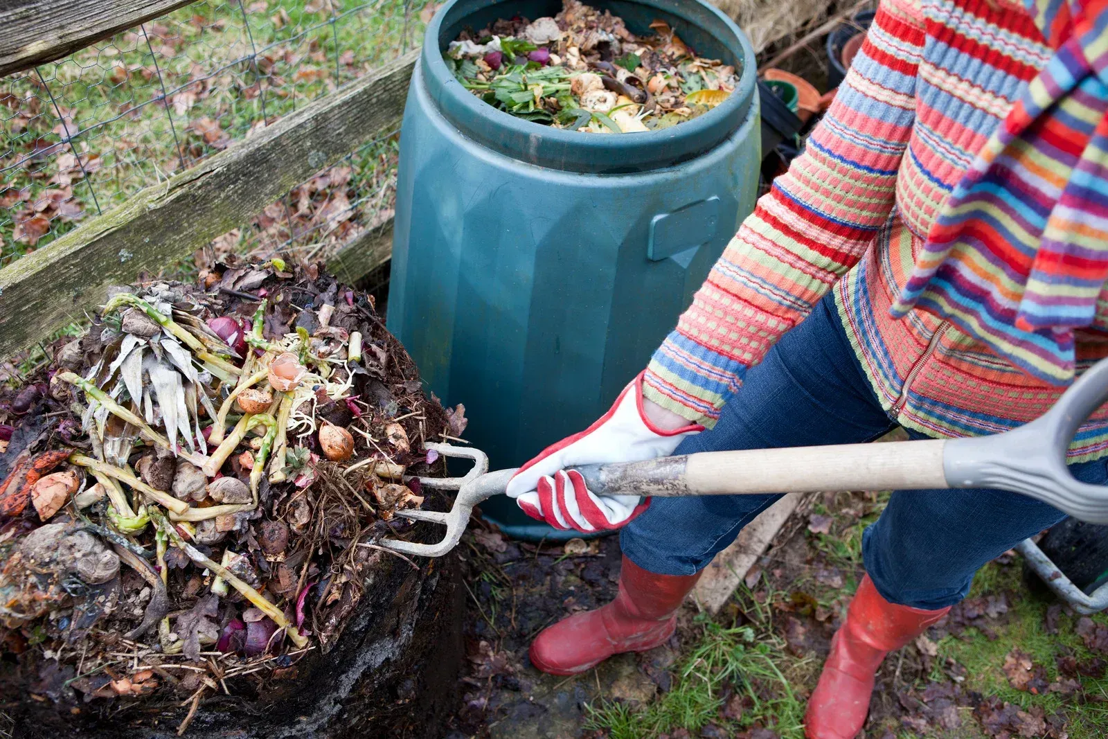 Woman in red boots uses a pitchfork to turn compost in a garden next to a green compost bin.