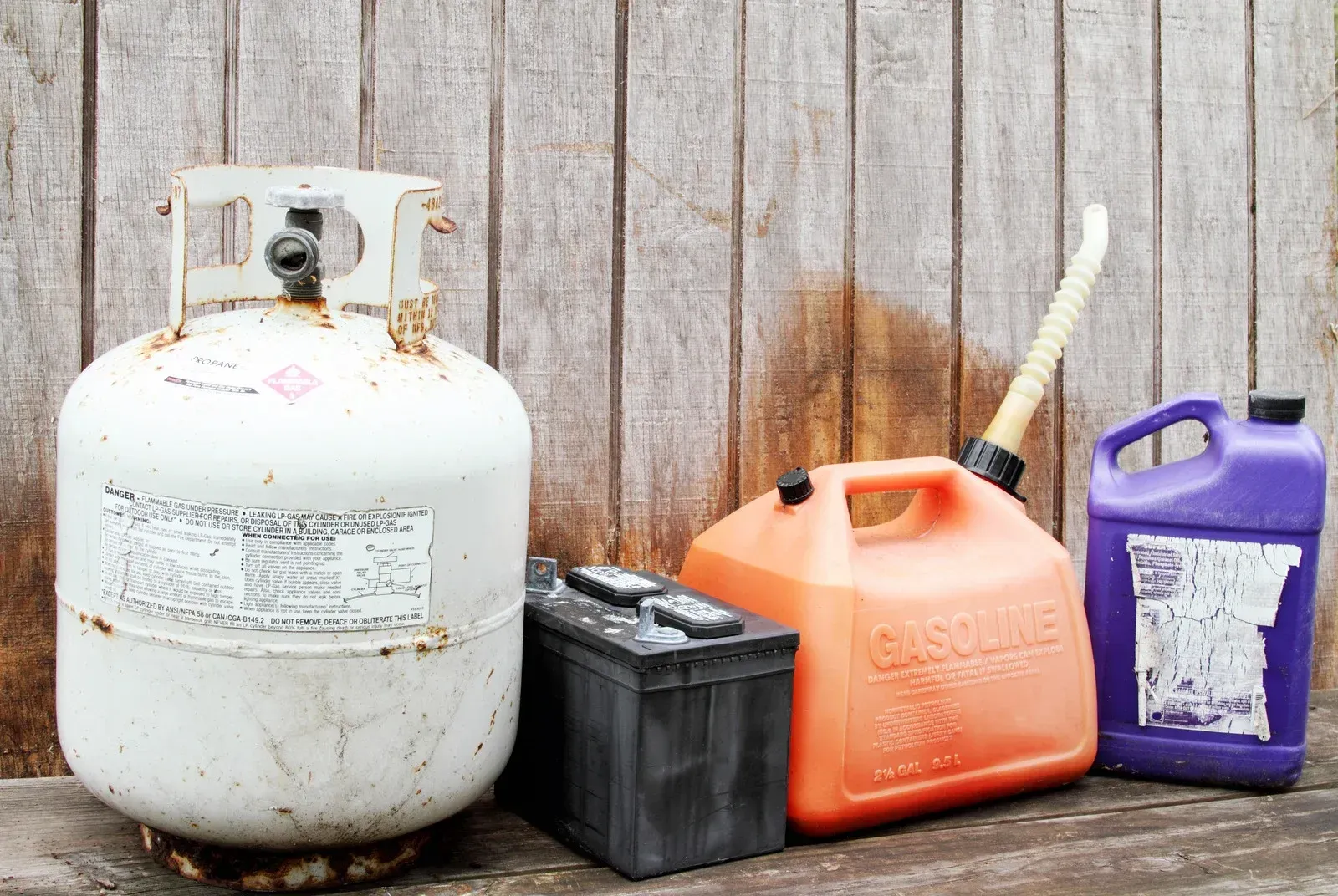 Propane tank, gas can, battery, and other containers against a wooden wall, representing potential hazards.