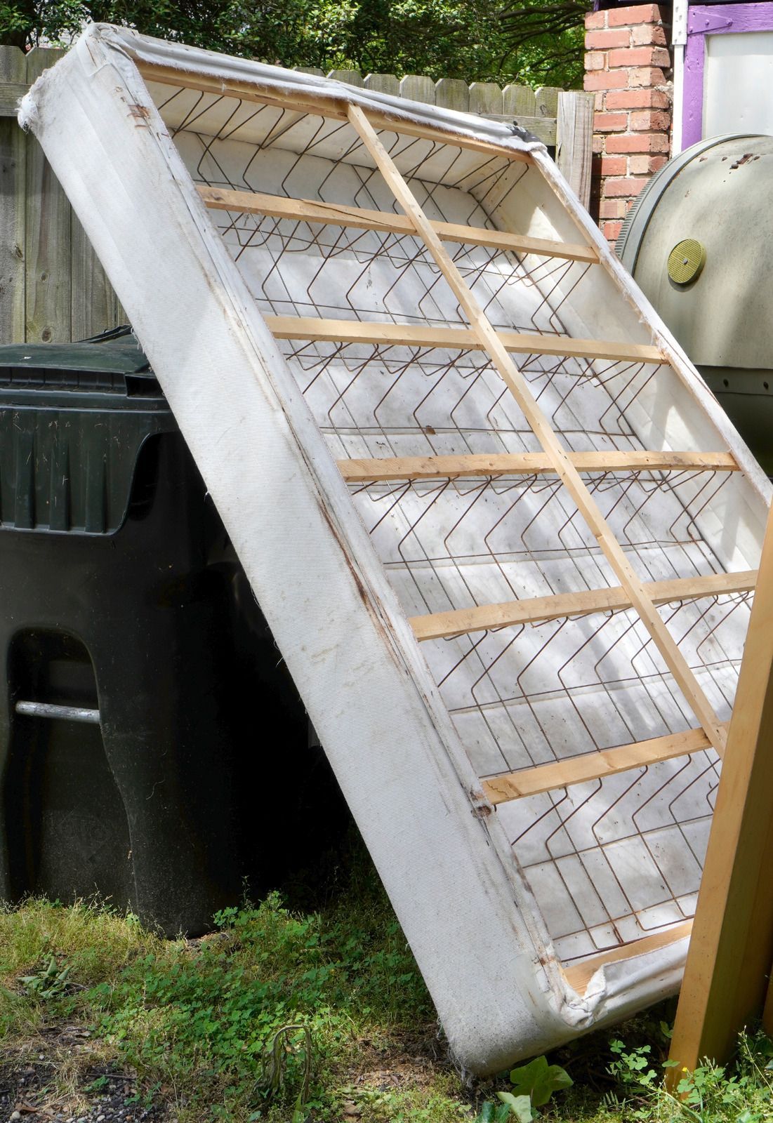 An old, white-upholstered mattress leaning against a black trash bin in a grassy yard.
