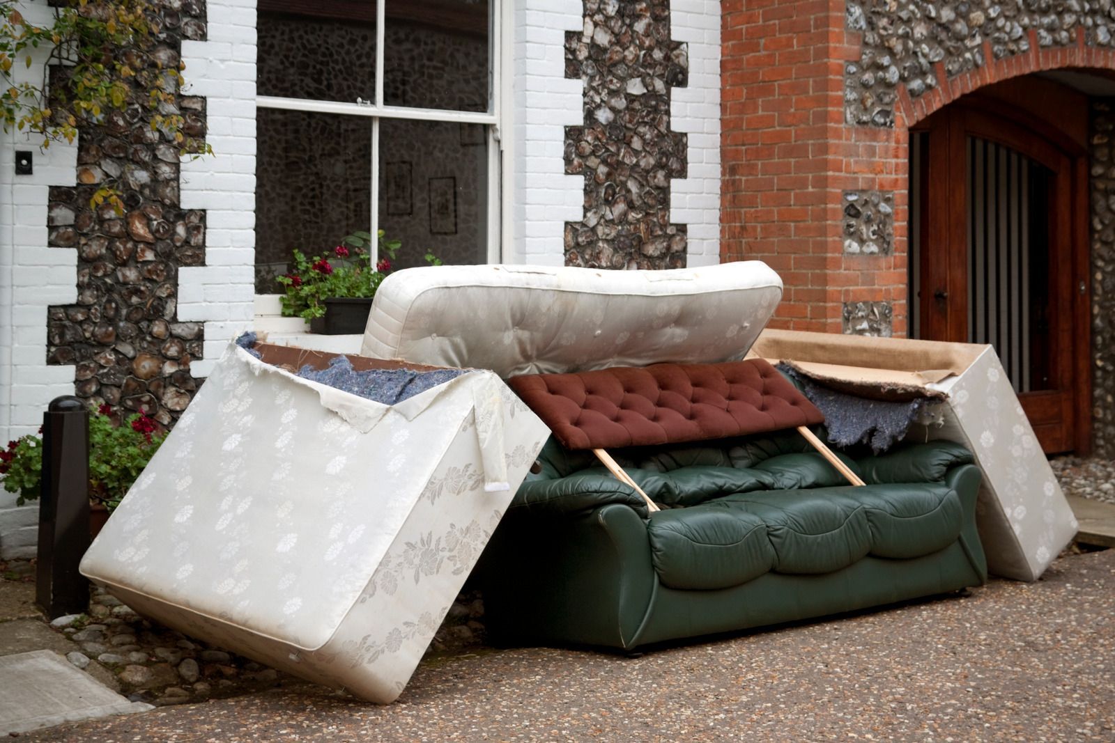 Discarded furniture pile: green couch, mattress, and cushions outside a brick building with a window.