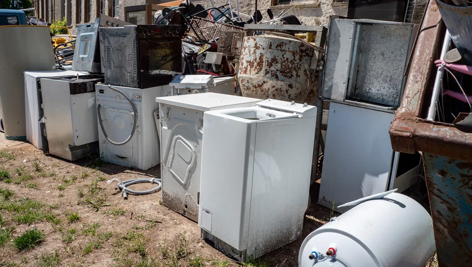 Heap of discarded white appliances, including washers and a water heater, outdoors.