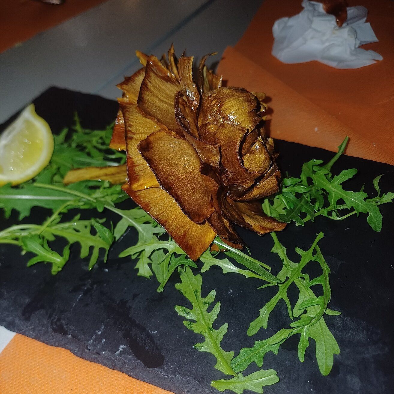 Pasta sfoglia fritta a forma di fiore, di colore dorato, servita su un'ardesia nera con verdure e uno spicchio di limone su un tavolo arancione.