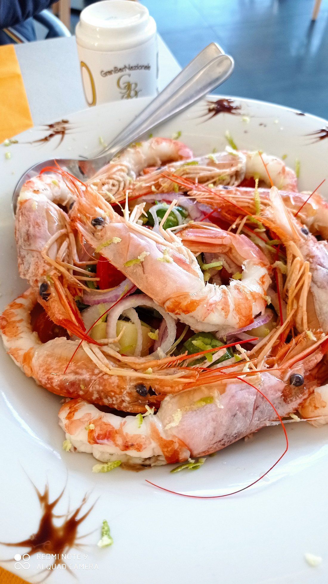 Un piatto di gamberi e spaghetti al sugo su un tavolo bianco, con una tazza e un tovagliolo sullo sfondo.