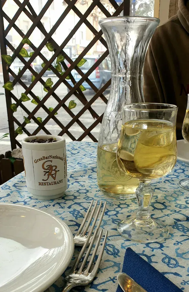 Tavolo da ristorante con piatti bianchi, forchette, una tovaglia blu a motivi geometrici e un bicchiere di vino bianco.