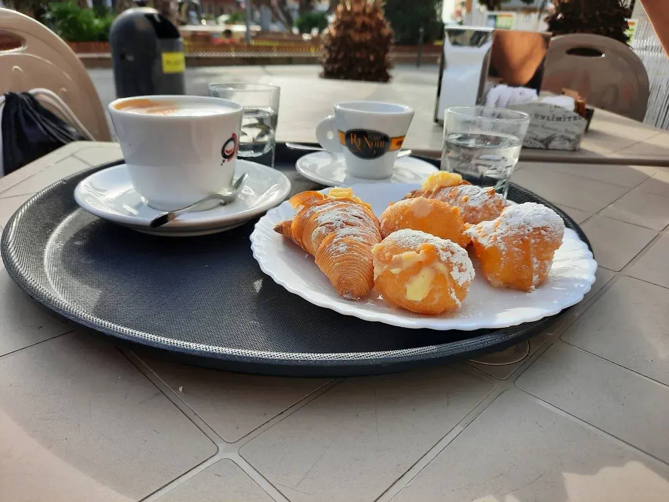 Tazza di caffè e pasticcini in polvere su un tavolino da bar all'aperto, con un bicchierino e un vassoio.