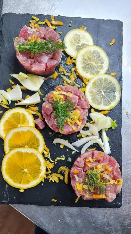 Tartare di tonno su tagliere di ardesia, guarnita con erbe aromatiche, fette di limone e scorza di agrumi.