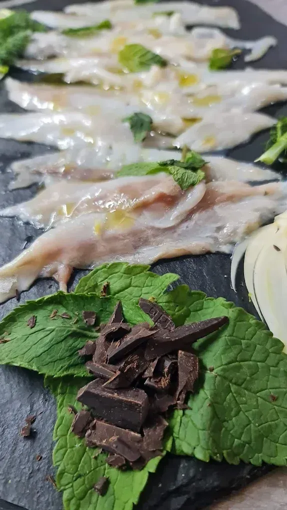 Fettine di pesce fresco e traslucido con foglie di menta e scaglie di cioccolato su un piatto di ardesia scura.