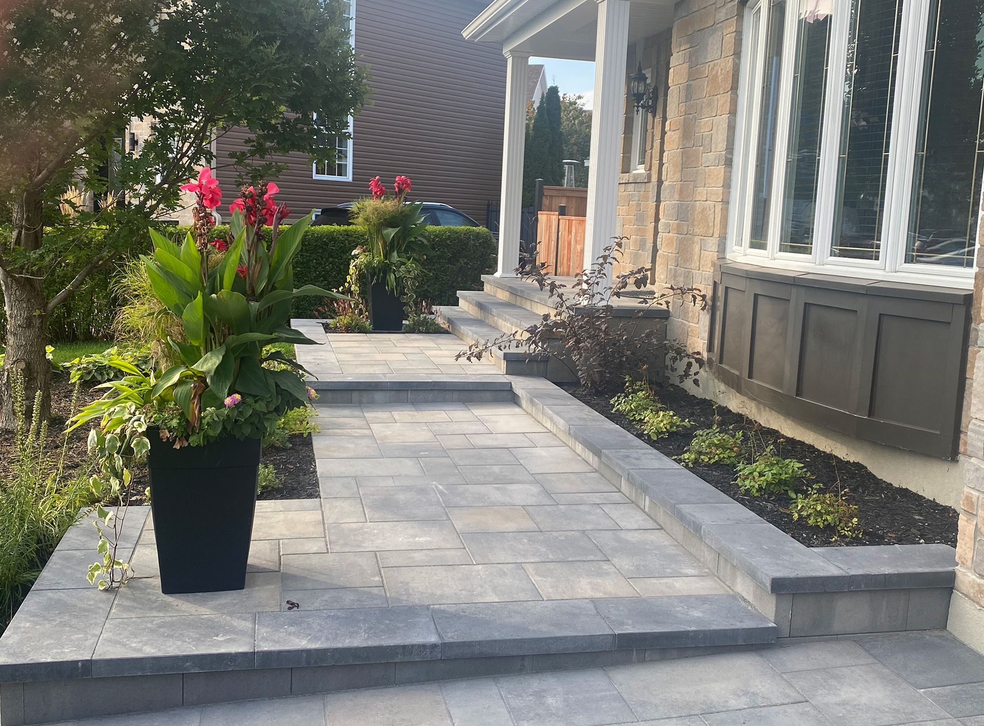 Allée pavée et marches menant à une maison à façade en briques. Des jardinières de fleurs rouges encadrent l'entrée.