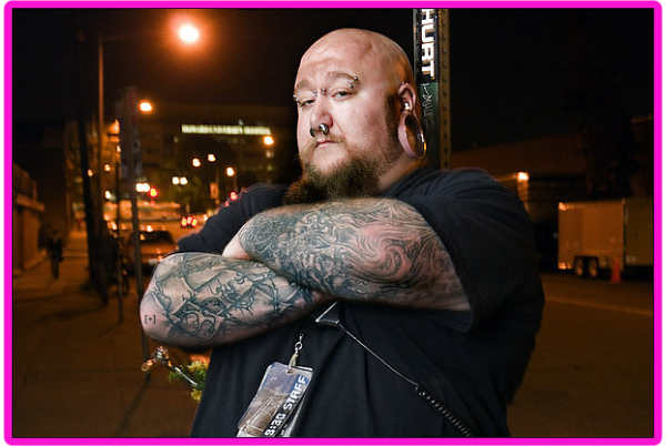 Man with tattoos and facial piercings stands outdoors at night, arms crossed, with a serious expression.