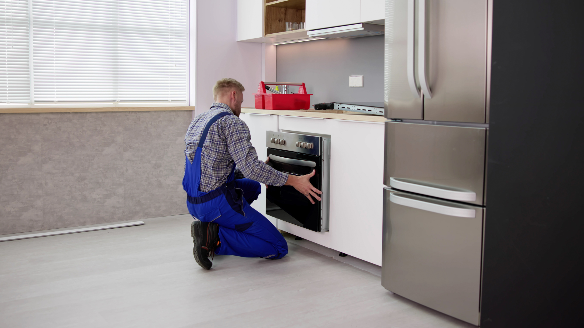 A repairman in blue overalls installs an oven in a modern kitchen with a stainless steel refrigerator.