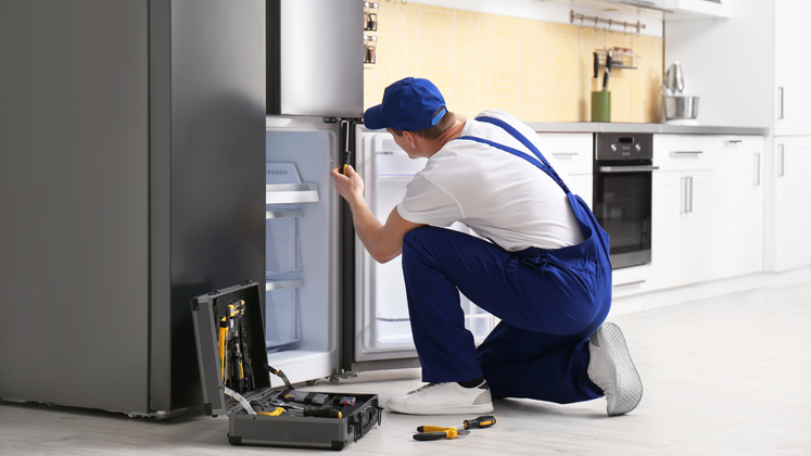 A repairman in blue overalls fixes a refrigerator in a kitchen. He is kneeling with a toolbox open nearby.