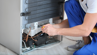 A person in blue overalls repairs the back of a refrigerator with a screwdriver.