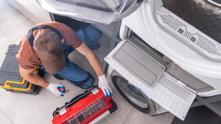 A repairman wearing gloves and a cap repairs a white washing machine, using tools from a red toolbox in a well-lit room.