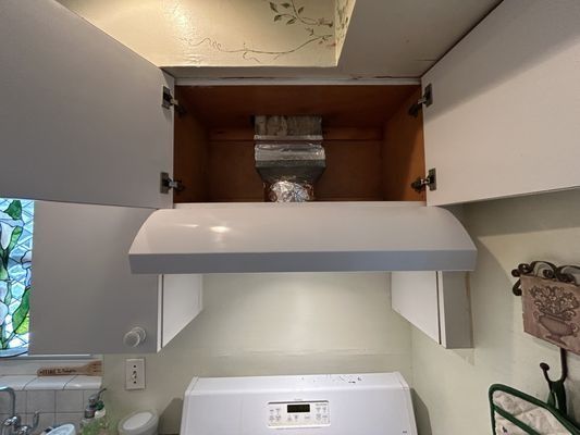 Kitchen range hood installed under white cabinets, with an exhaust vent visible. The cabinets are open, and the stove is below.