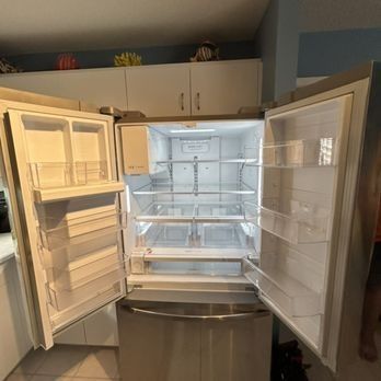 A stainless steel refrigerator with all doors open in a kitchen. Interior shelves and bins are visible and empty.