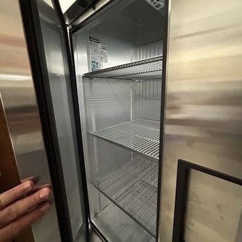 An empty stainless steel refrigerator with wire shelves, open revealing the interior. A hand reaches in to hold the door.
