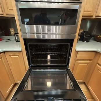 An open stainless steel oven with a microwave above, built into light wood cabinets.