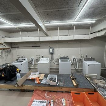 Four washing machines lined up against a wall in a utility or service room, with work tables in front.