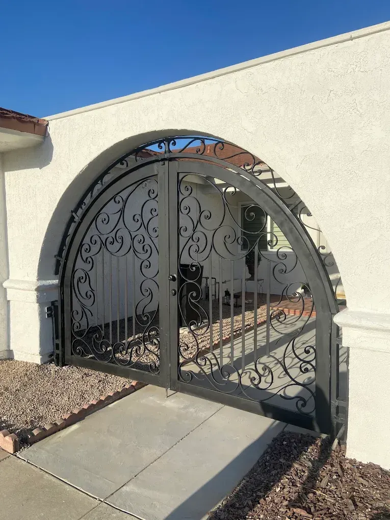 Black wrought iron gate set within an arched entryway. The gate is in front of a pathway with rocks and light-colored walls.