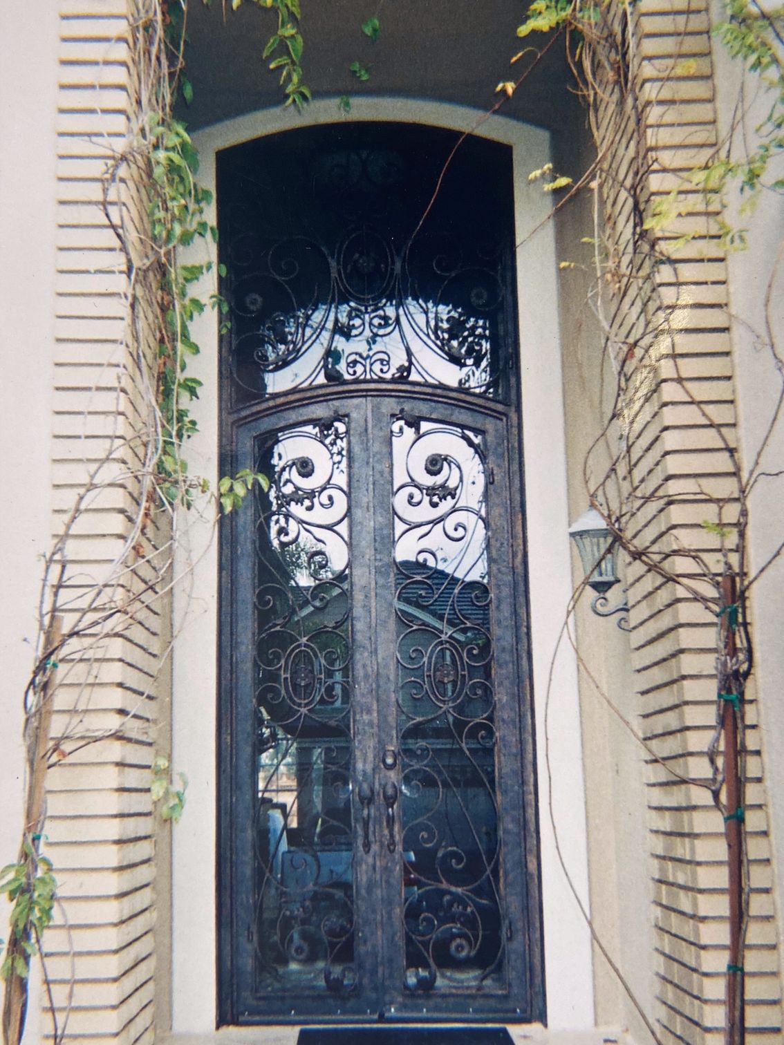 Ornate wrought-iron double doors with arched top, framed by brick pillars and climbing vines.