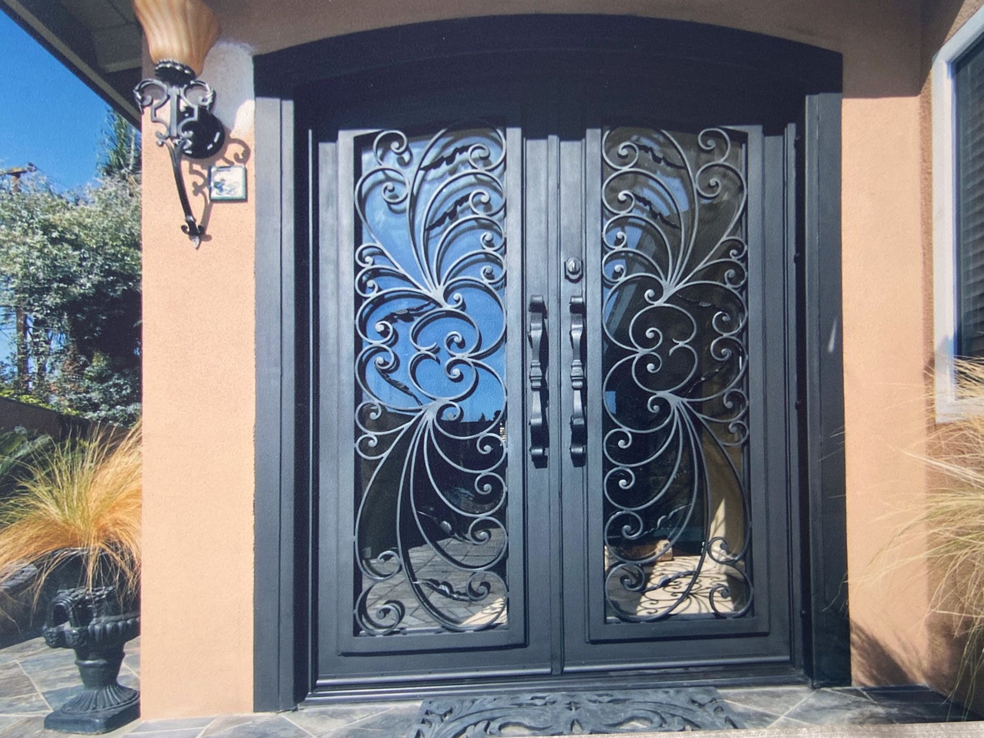 Black wrought-iron double doors with ornate designs; set in a stucco exterior, flanked by plants and a sconce.