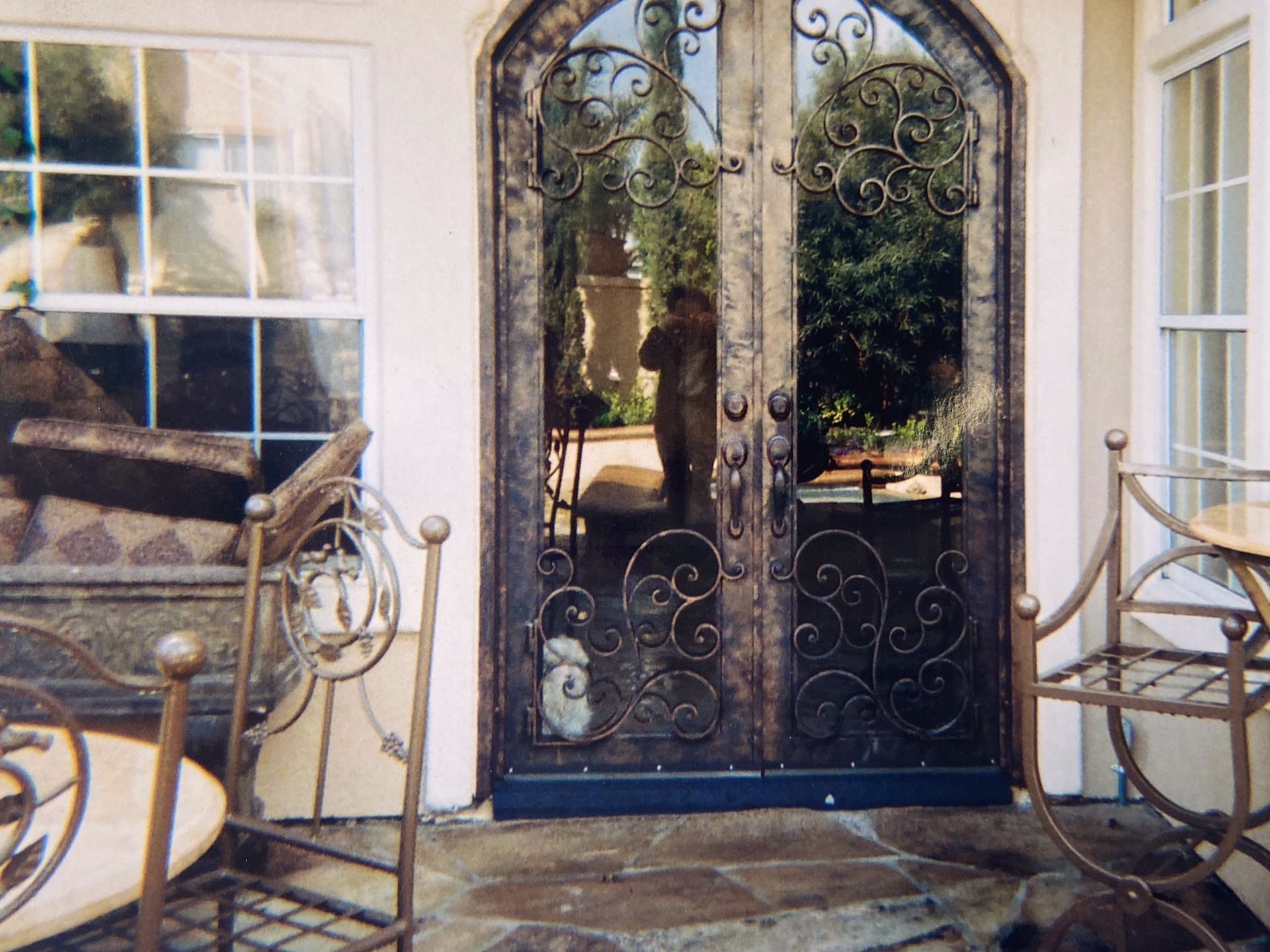 Ornate wrought iron double doors reflecting outdoor scene and person. Chairs and window on either side.