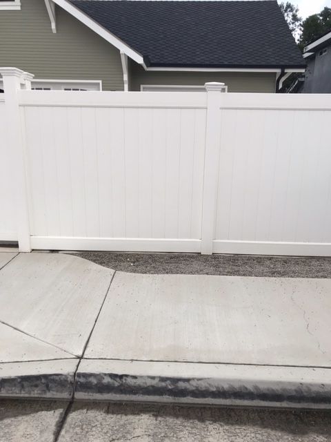 White fence along a sidewalk. Gray gravel below the fence. Green house in the background.