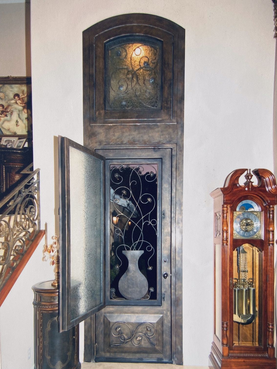 Ornate iron door with open side panel revealing a stained glass design. Above is a matching arched window. A tall grandfather clock stands to the right.