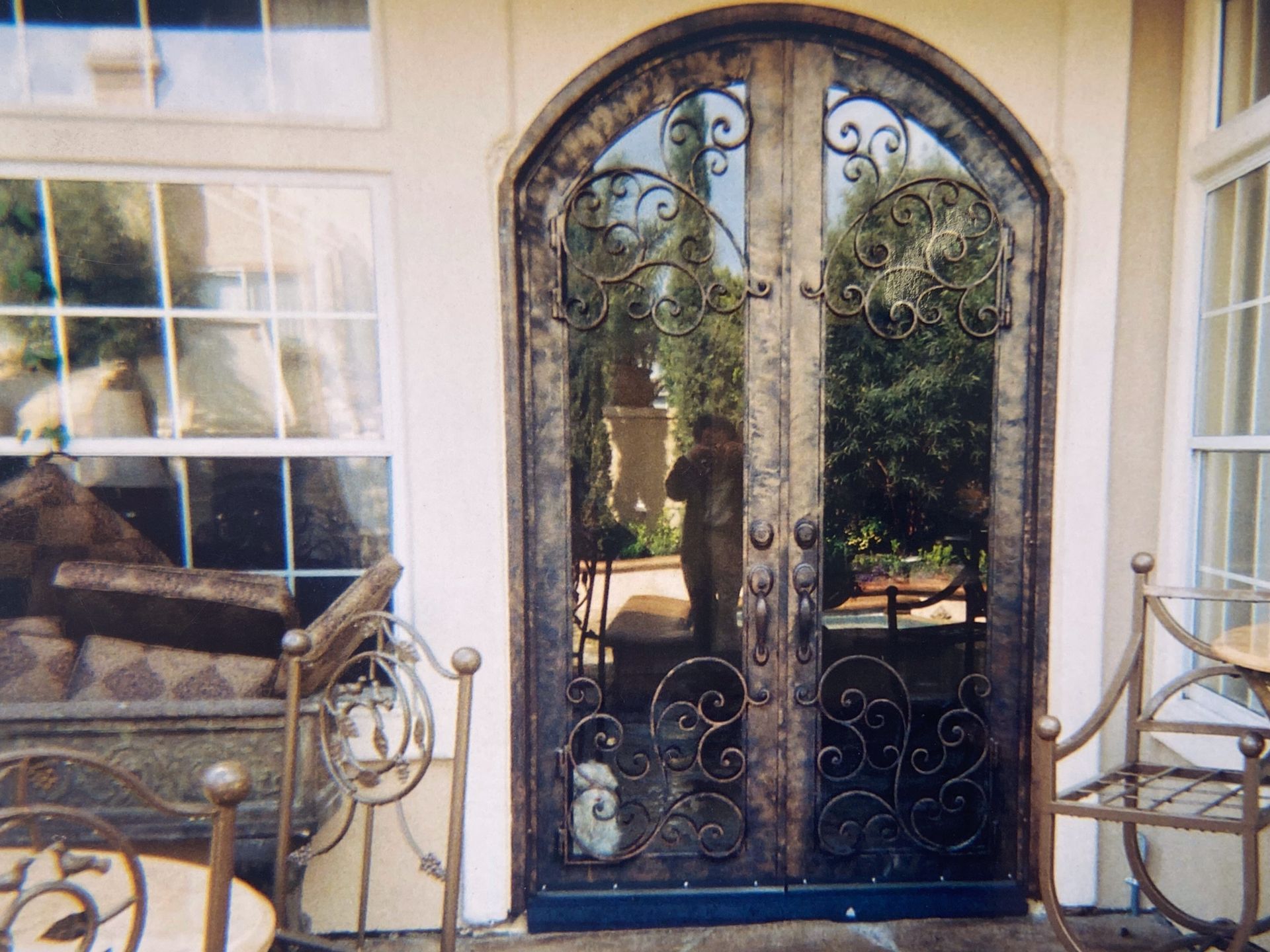 Ornate, arched double doors with wrought iron design and glass panels reflect a garden scene.