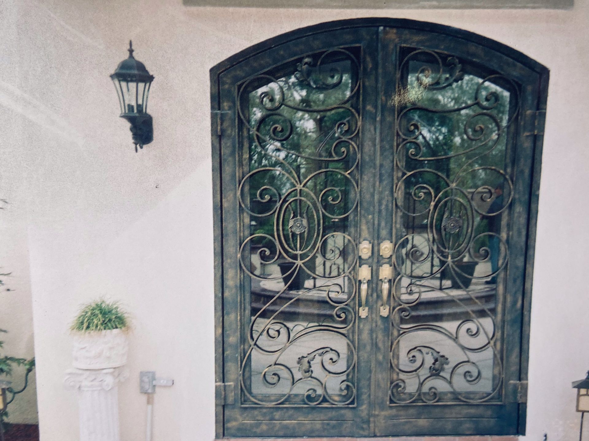 Ornate wrought iron double doors with glass panels, a wall-mounted lamp, and a potted plant.