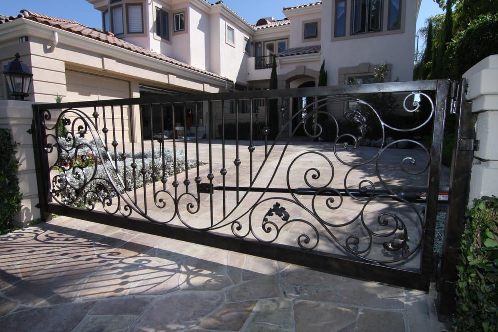 Black wrought-iron driveway gate with decorative scrollwork in front of a large house.