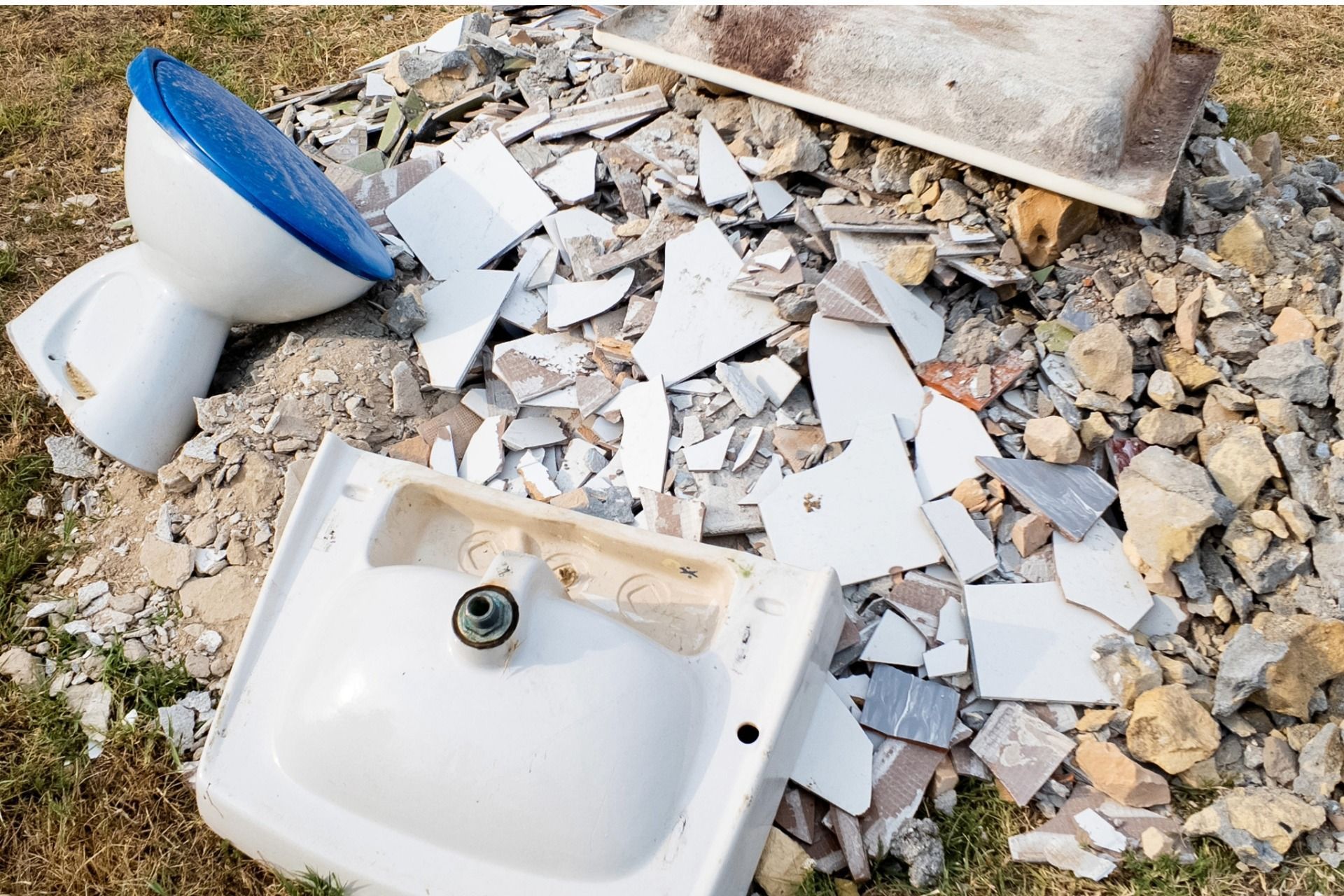Pile of broken bathroom fixtures: toilet, sink, tub, and tile rubble outdoors.