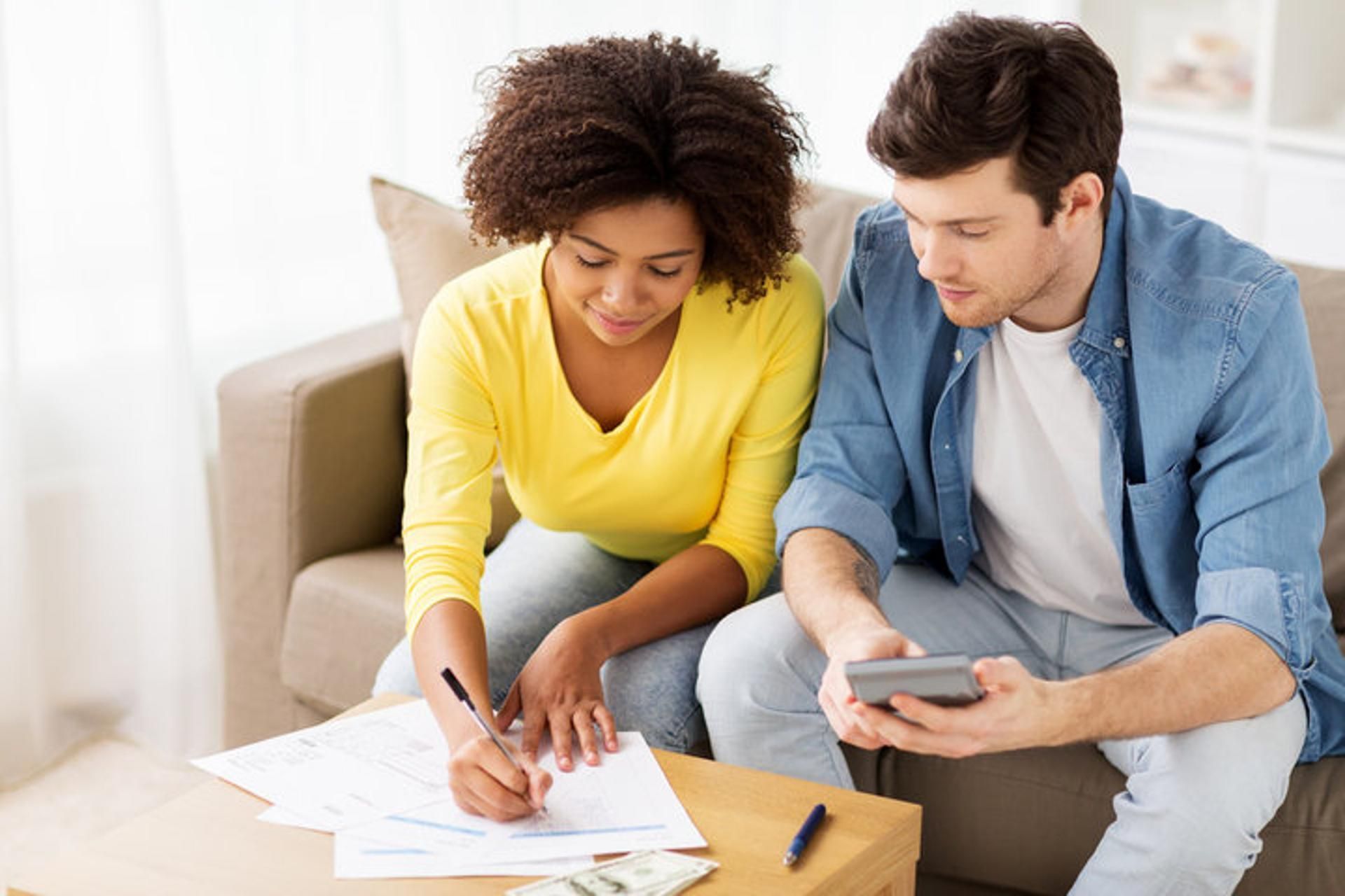 Two people sitting on a couch, reviewing documents and using a calculator to manage their personal finances.