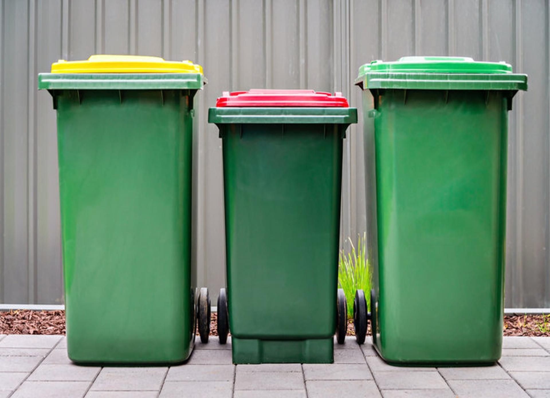 Green trash bins overflowing with black garbage bags against a blurred building background.