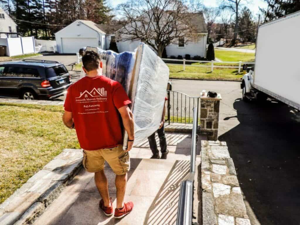 bedroom furniture is being moved out from the house carefully using the stairs. we see a black SUV on the left of the frame and a white moving truck on the far right.