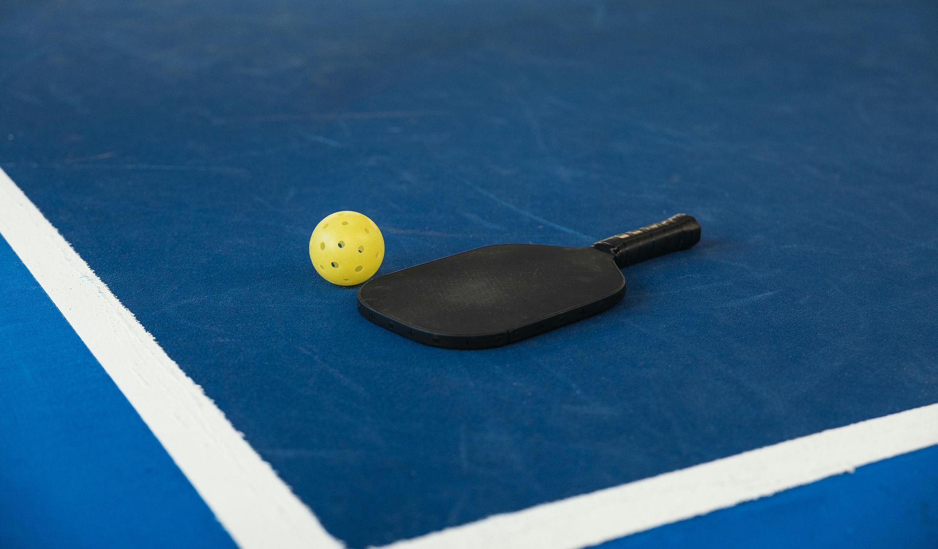 Black pickleball paddle and yellow ball on a blue court next to a white line.