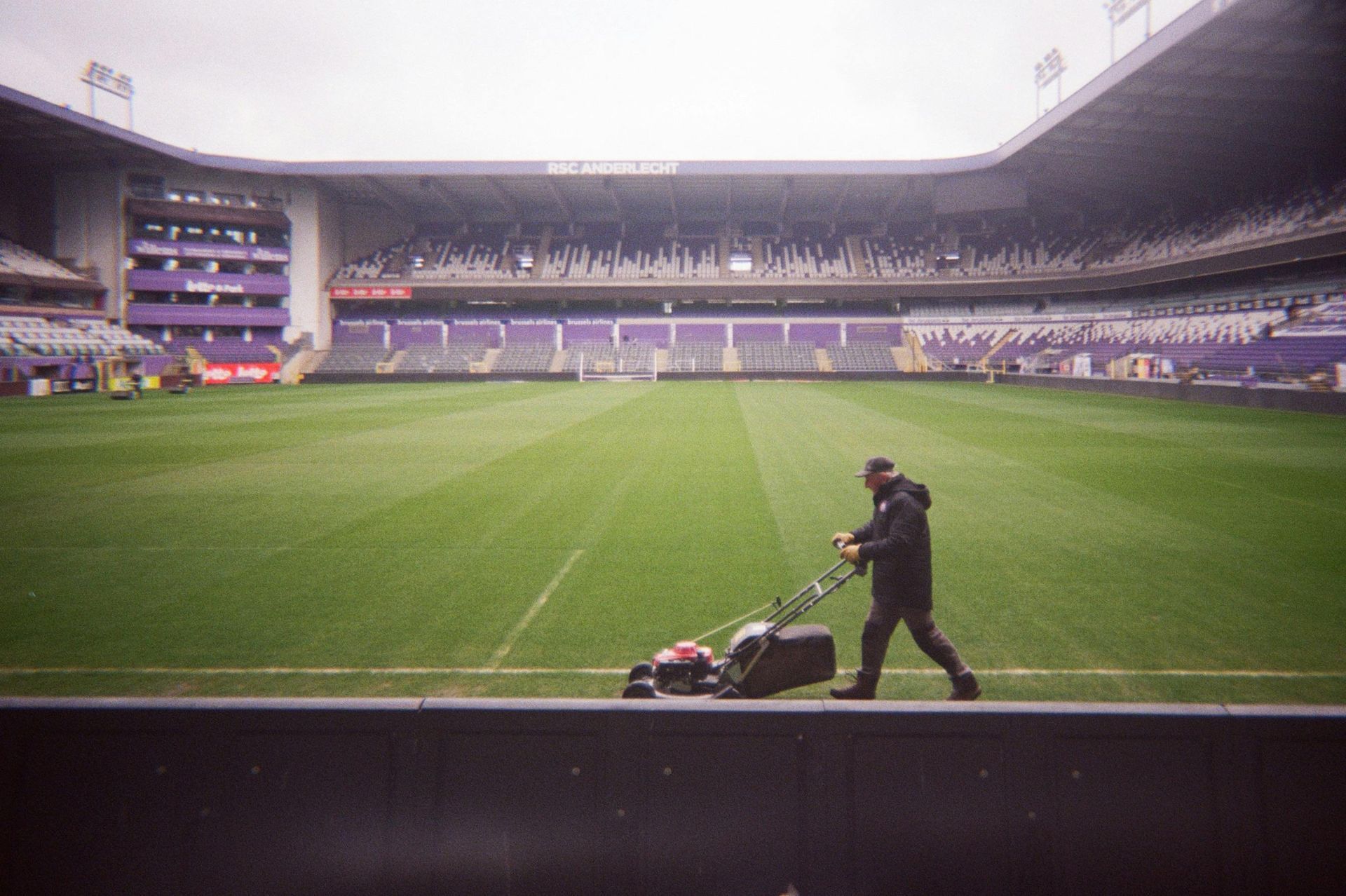 Man mowing the grass on a soccer field in a stadium with purple seating.