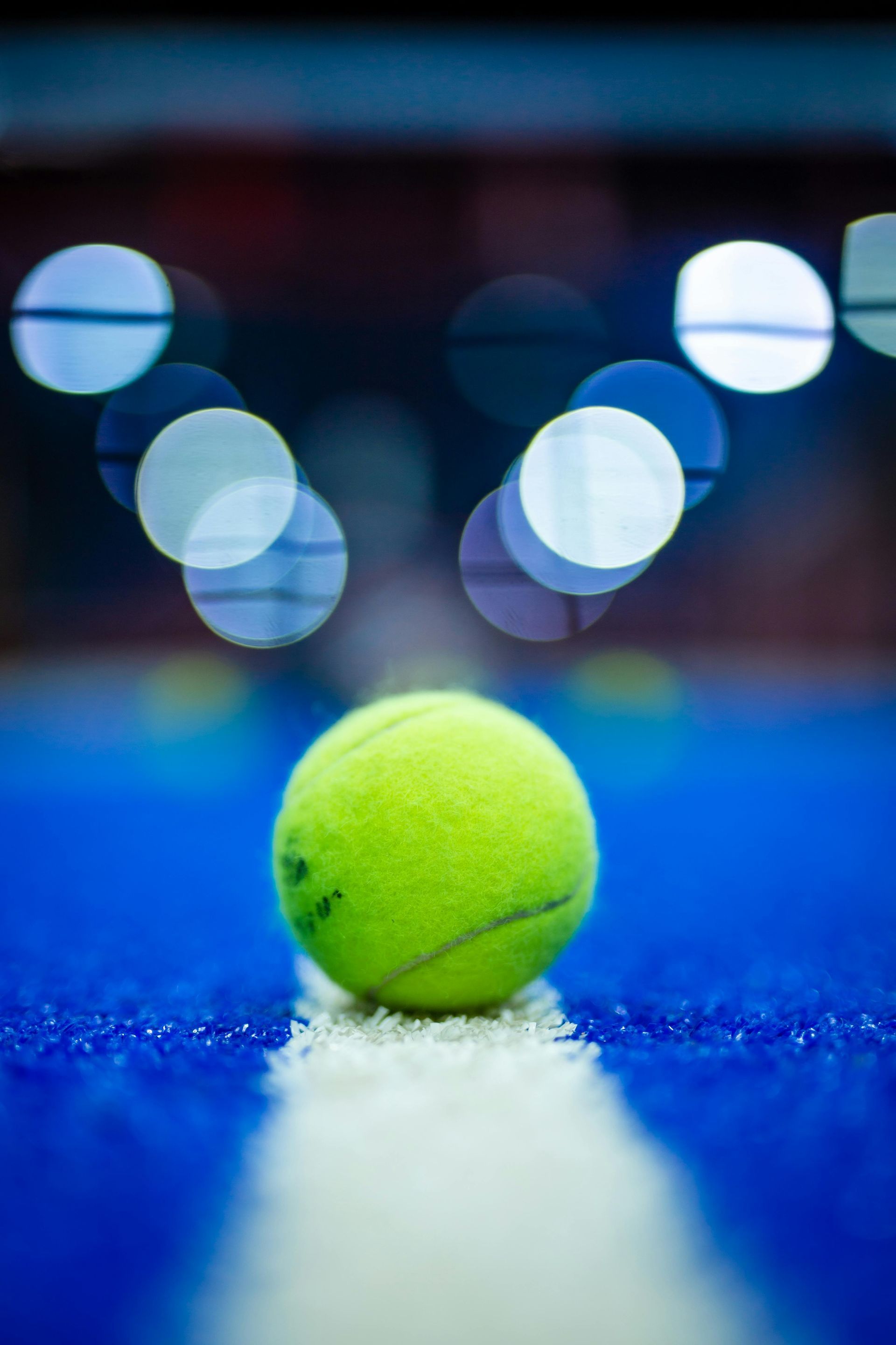 Tennis ball on a white line on a blue court, with blurred lights in the background.
