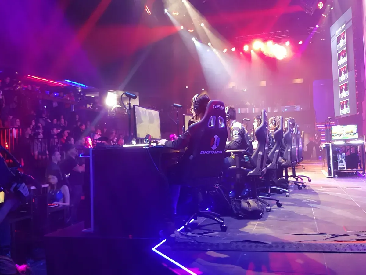 Esports stage with players at PCs under red and purple lights, audience watching from the side.