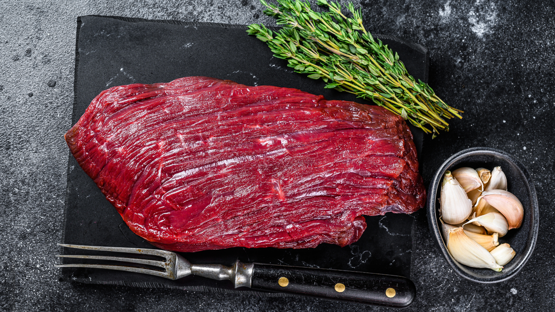 A piece of raw beef steak on a cutting board with garlic and thyme.