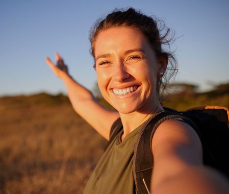Smiling hiker taking a selfie at sunset on a grassy hill, arm outstretched.