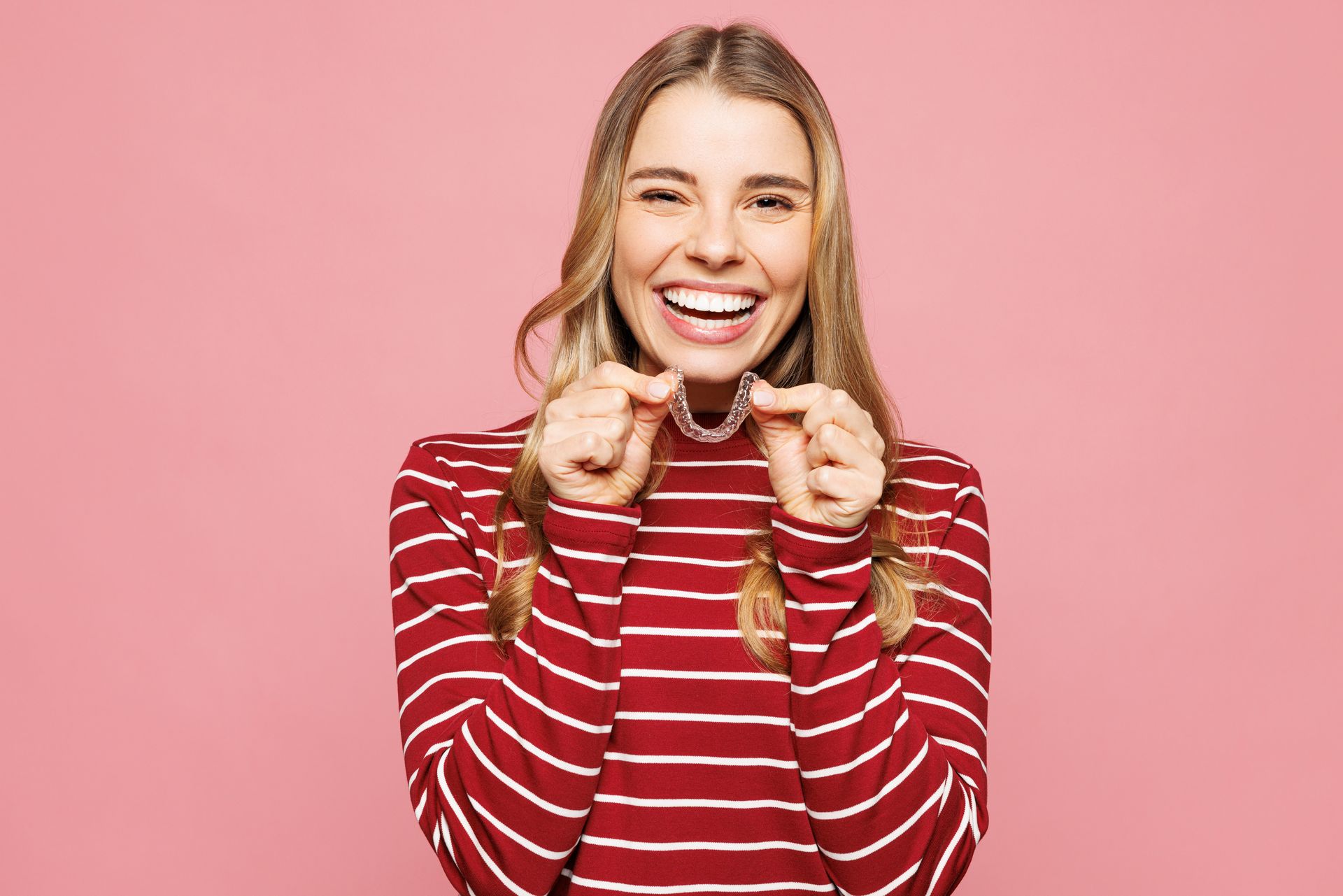 Woman smiling, holding clear dental retainer, pink background.