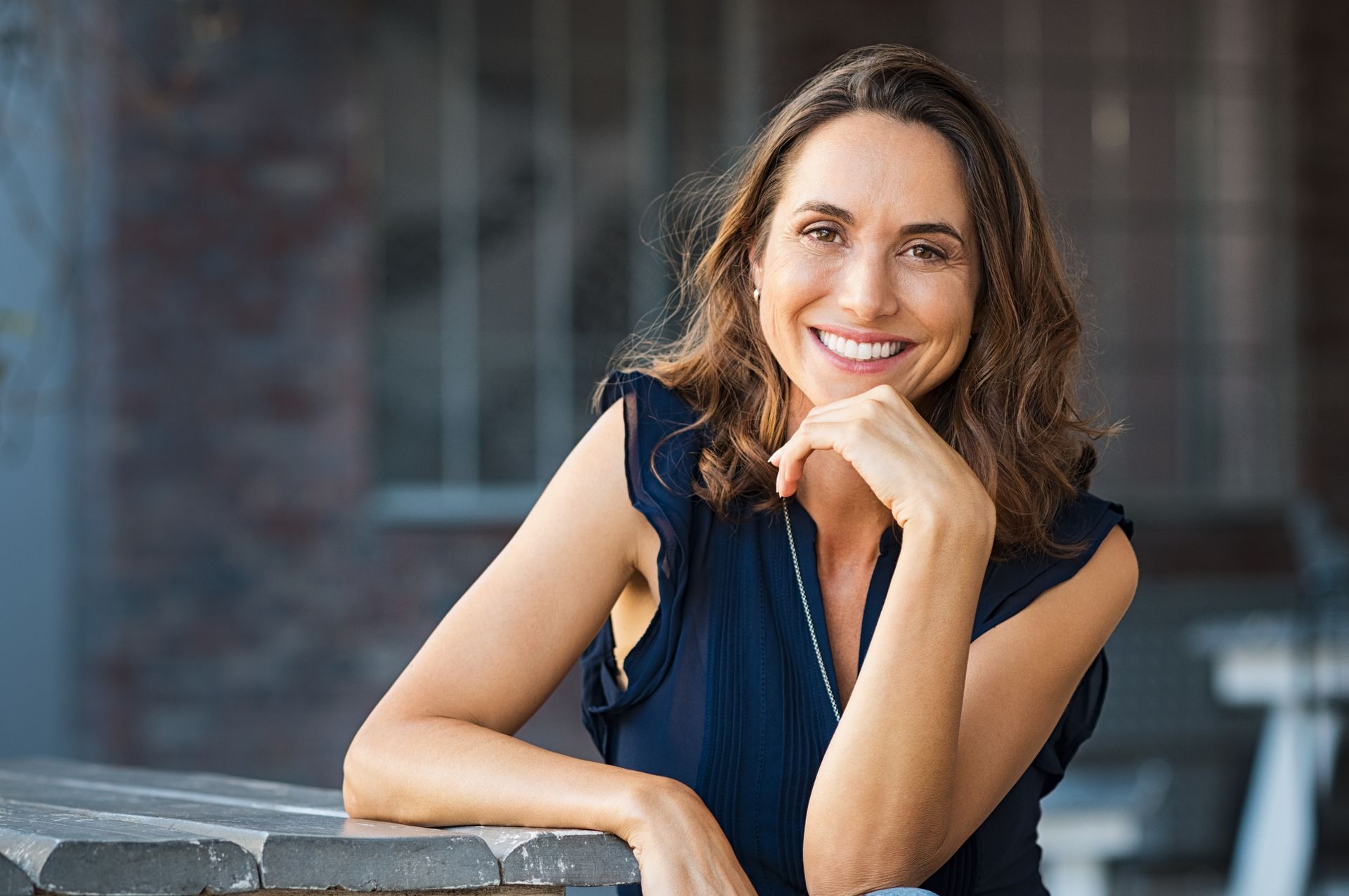 Woman in a navy top smiles, resting chin on hand. Leans on a weathered gray table in an outdoor setting.