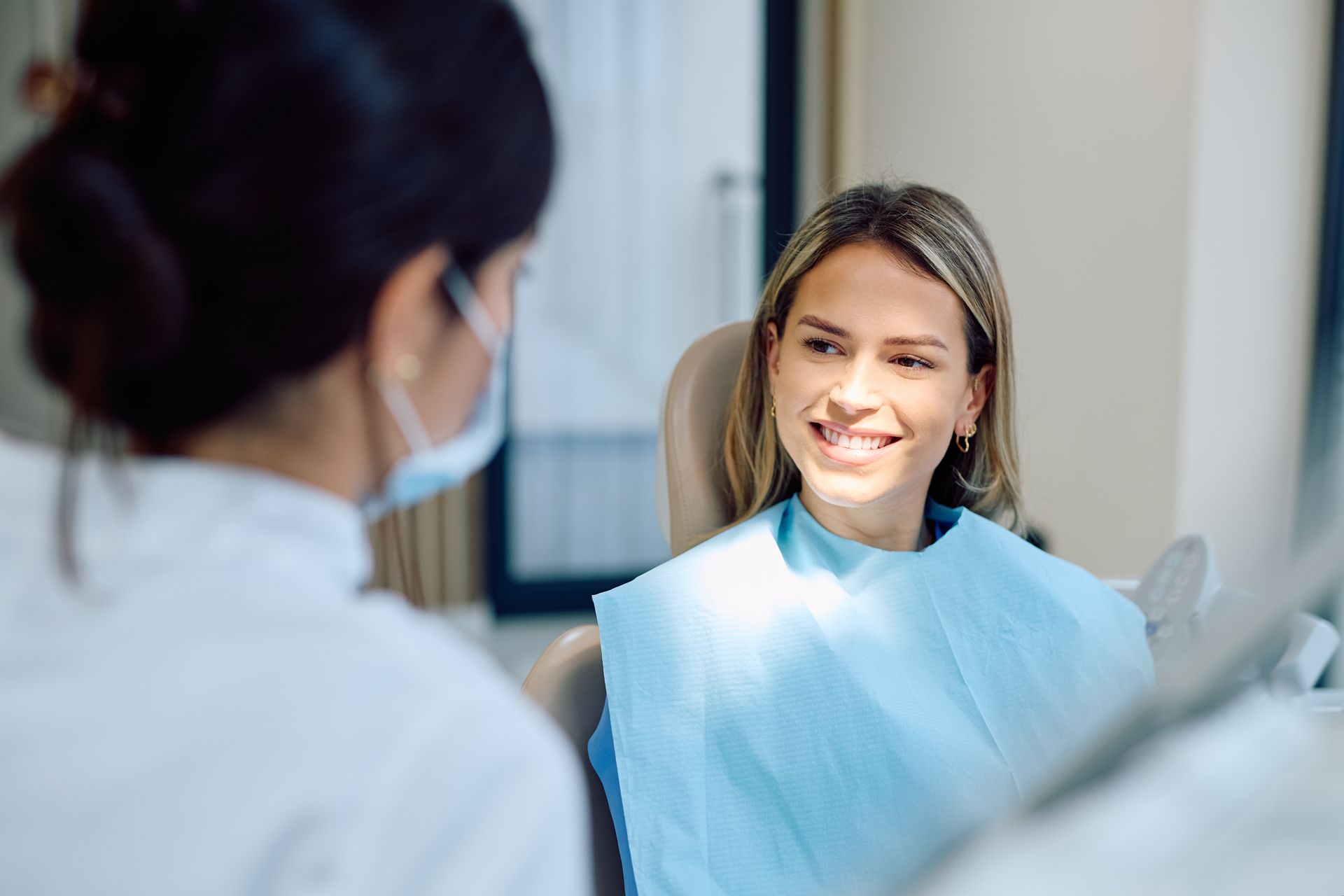 Patient smiling at a dentist during a dental exam, wearing a blue bib in a bright clinic.
