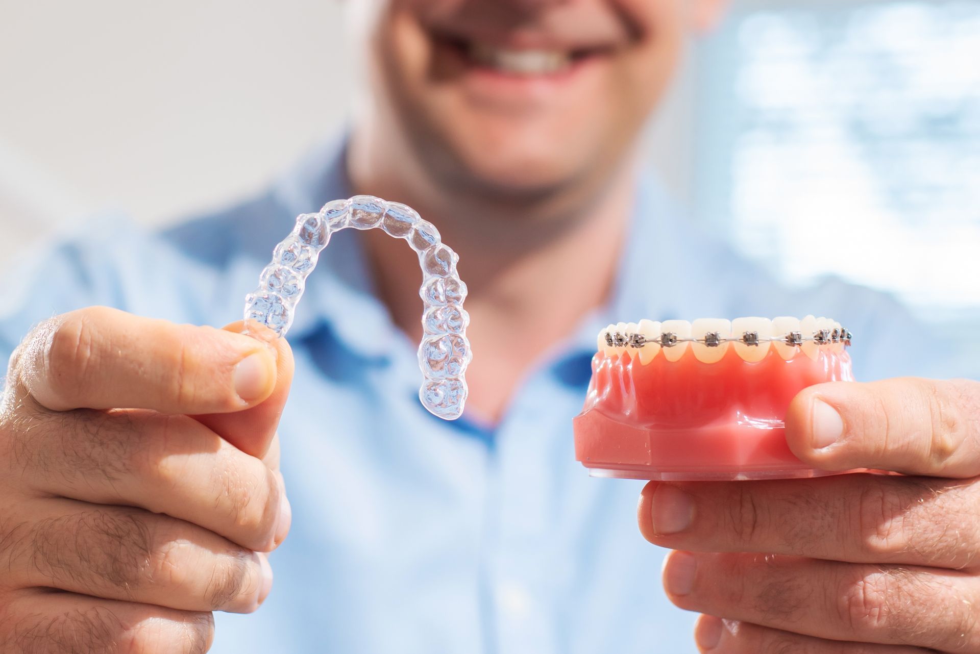 Person holding clear aligner and a model with braces, smiling.