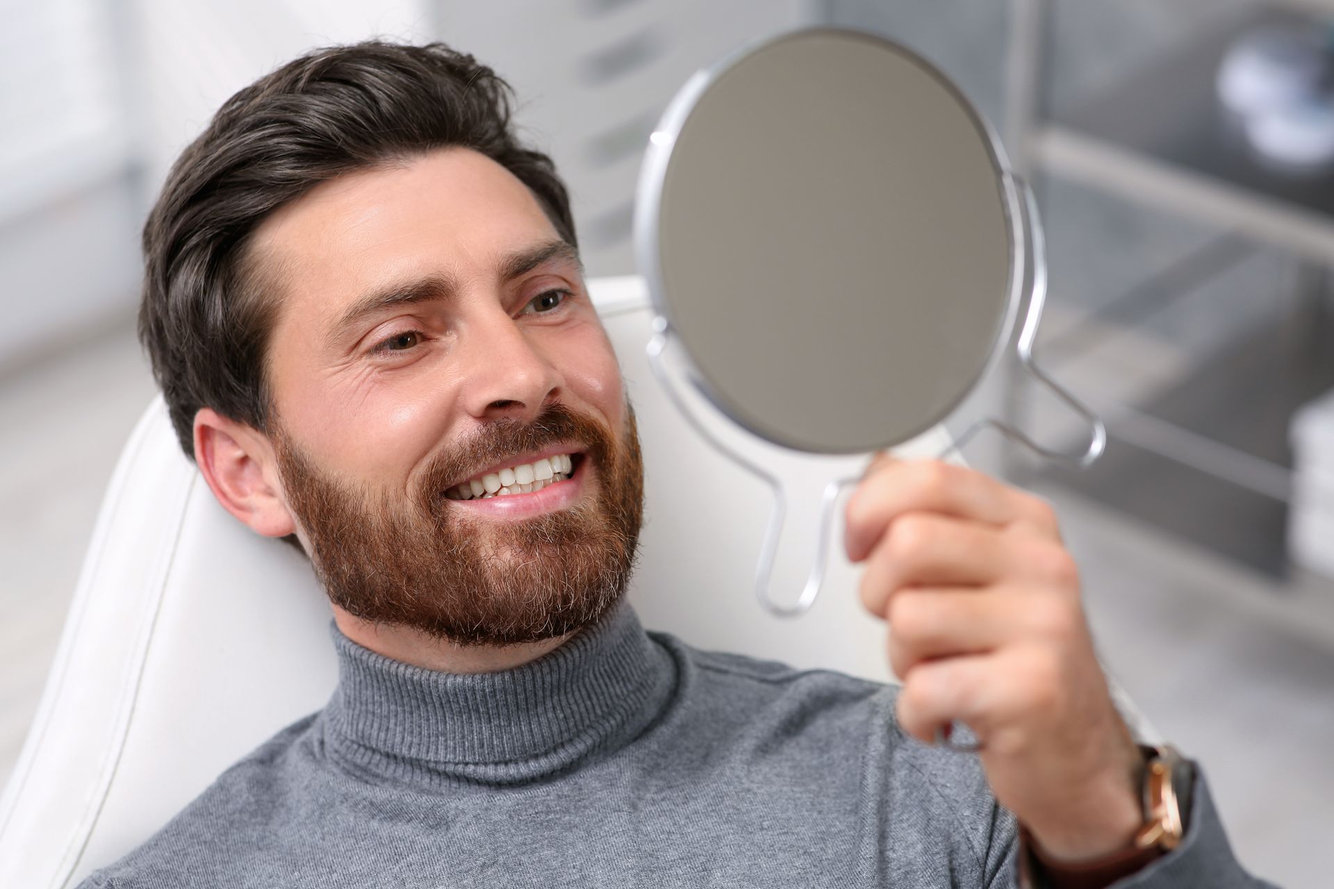 Man smiling at his teeth in a handheld mirror, seated in a dental chair.
