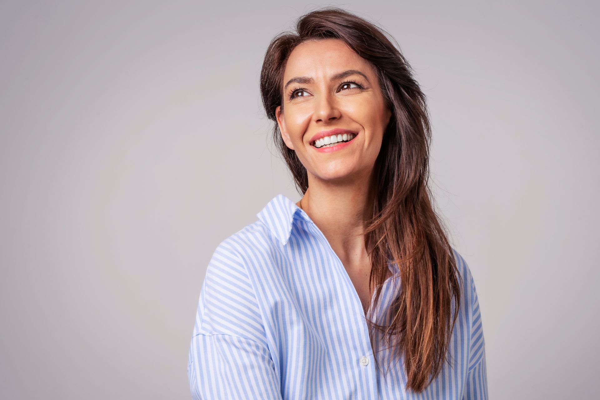 Woman smiling, looking upward. Wearing a light blue striped button-down shirt, against a light gray background.