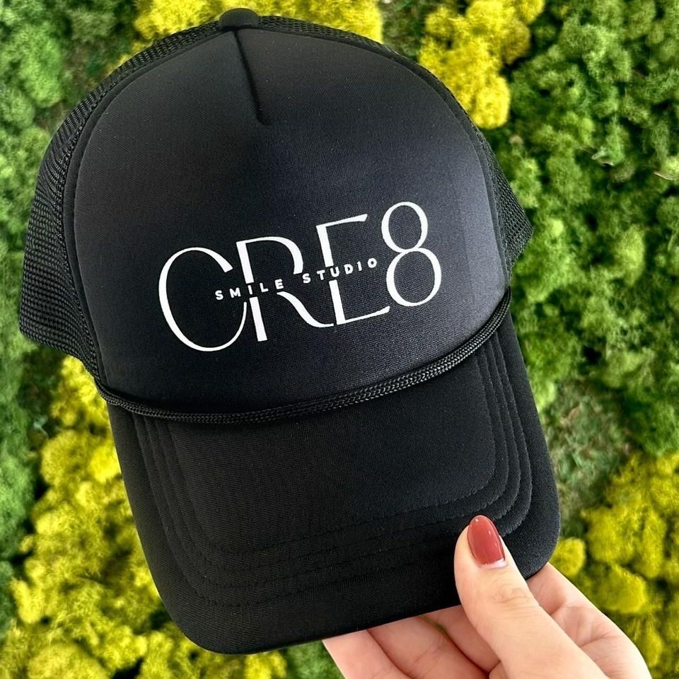 A person is holding a black hat that says cre8 smile studio
