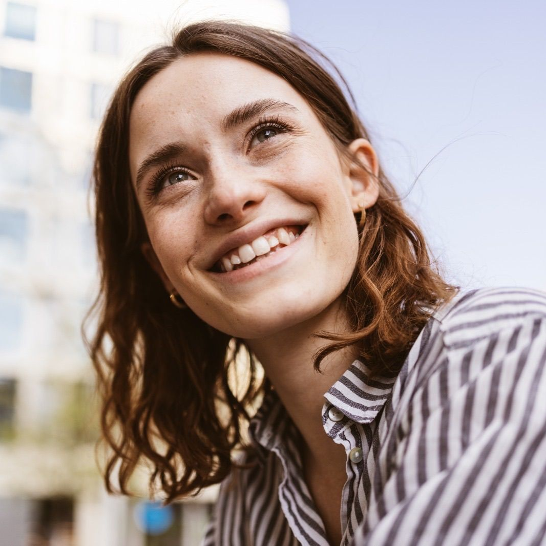 Woman with brown hair looking upward, smiling. Wearing a striped shirt, outside near a building.